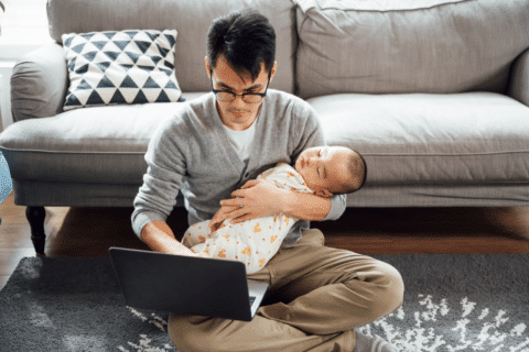 Young Asian father holding his little cute sleeping baby son while working with computer at home.