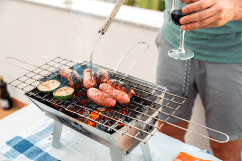 From above of crop anonymous man in casual clothes preparing sausages and zucchini slices on portable BBQ grill and drinking glass of red wine standing on terrace at home